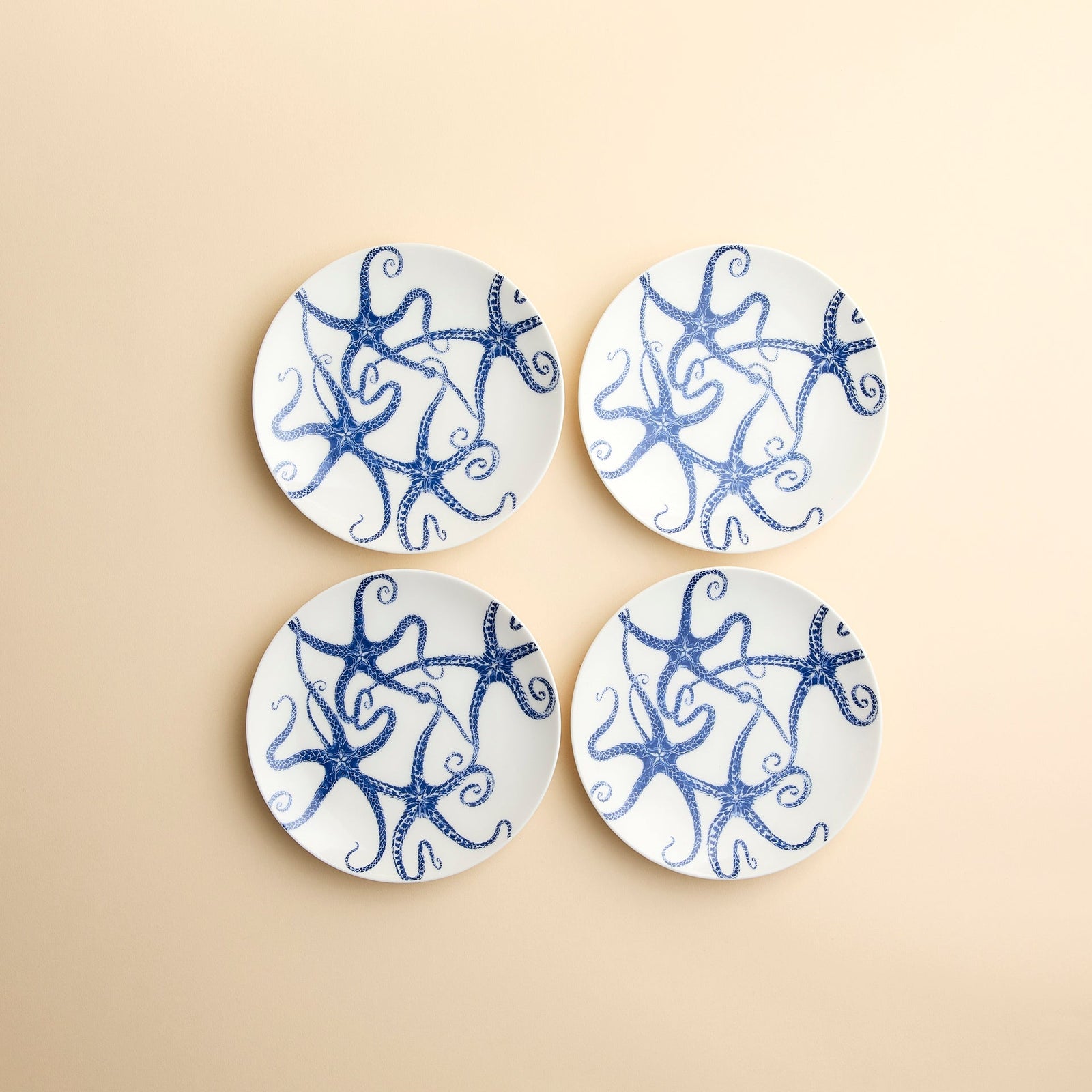 Whimsical Starfish dance across the faces of this set of 4 small appetizer plates from Caskata in creamy blue and white porcelain against a beige background.