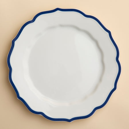 The Stella Scalloped Blue Dinner Plate by Caskata, seen from above, emanates a vintage charm with its wavy blue rim, offering an heirloom-quality dinnerware aesthetic on a creamy beige background.