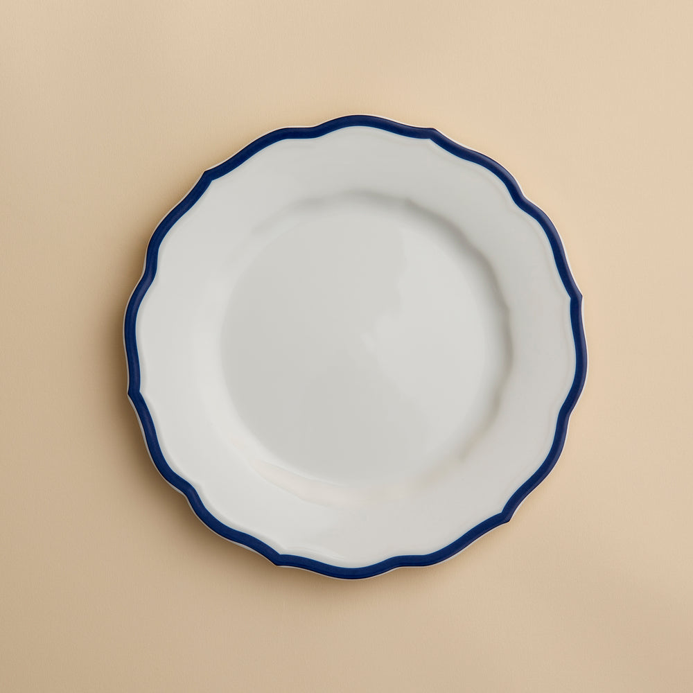 A white porcelain salad plate with a wavy, dark blue rim—the Stella Scalloped Blue Salad Plate by Caskata—is centered on a light beige background. This plate combines elegance and practicality, as it is both microwave and dishwasher safe.