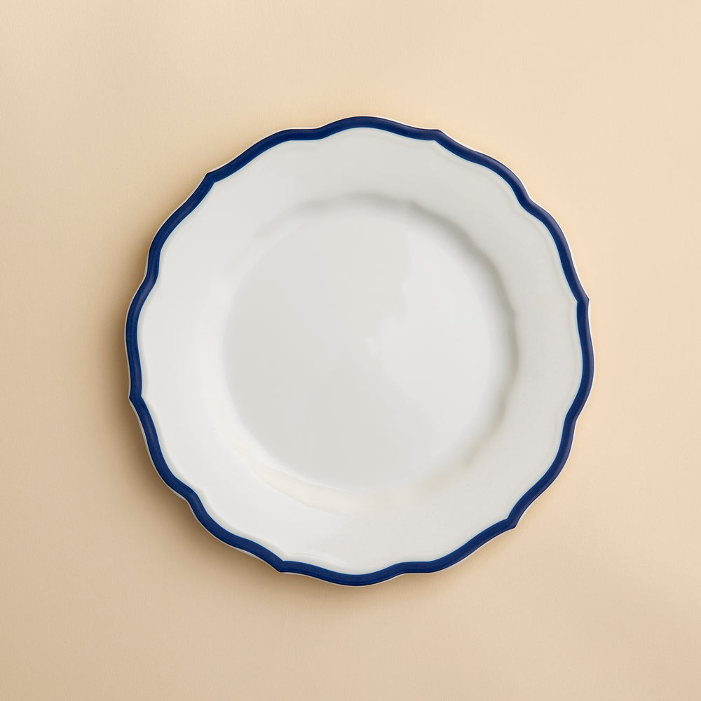 A white porcelain salad plate with a wavy, dark blue rim—the Stella Scalloped Blue Salad Plate by Caskata—is centered on a light beige background. This plate combines elegance and practicality, as it is both microwave and dishwasher safe.