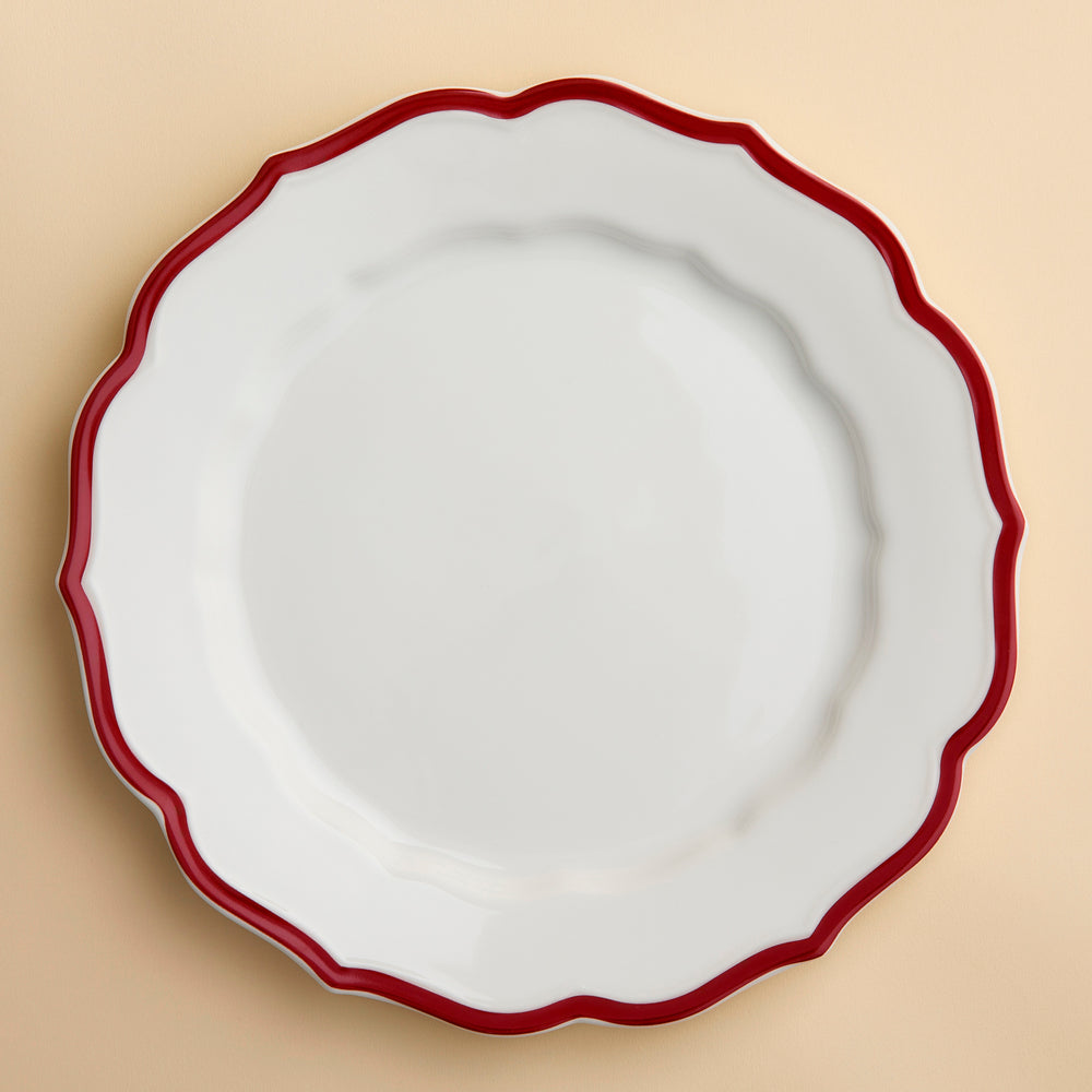 The Stella Scalloped Crimson Dinner Plate by Caskata is an heirloom-quality, high-fired porcelain plate with a scalloped edge and a bold red border, shown from above against a creamy beige background.