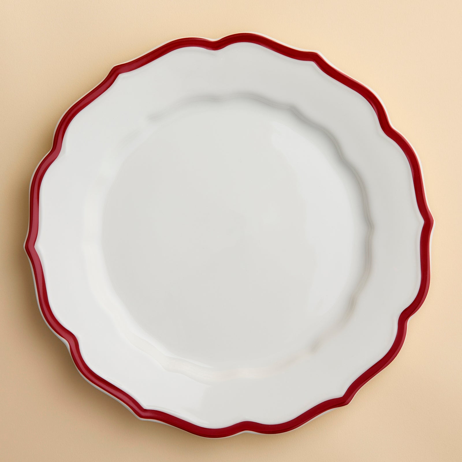 The Stella Scalloped Crimson Dinner Plate by Caskata is an heirloom-quality, high-fired porcelain plate with a scalloped edge and a bold red border, shown from above against a creamy beige background.