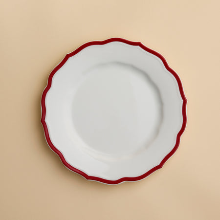 The Stella Scalloped Crimson Salad Plate by Caskata, featuring a white ceramic surface with a wavy crimson rim inspired by vintage dinnerware, is displayed on a light beige background.