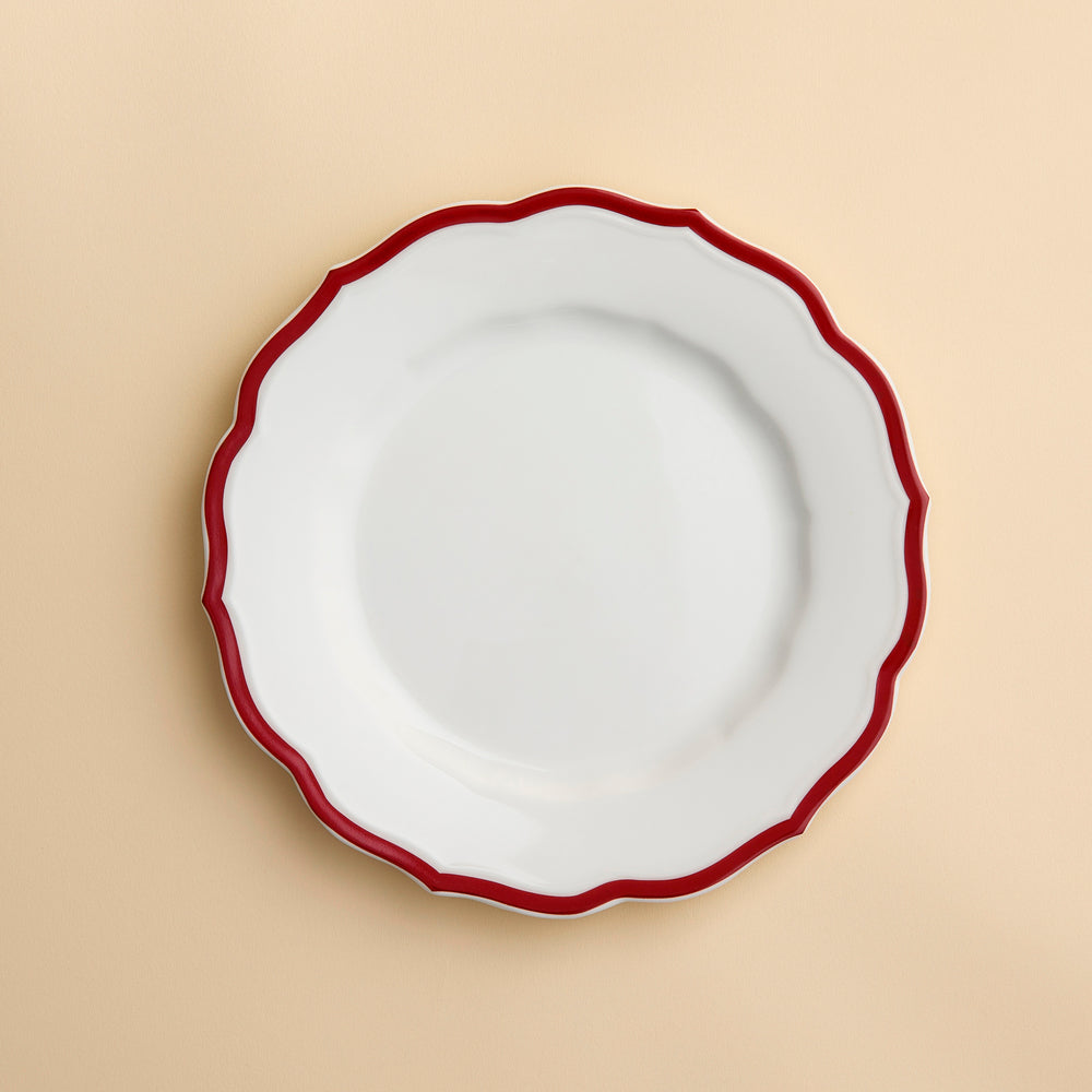 The Stella Scalloped Crimson Salad Plate by Caskata, featuring a white ceramic surface with a wavy crimson rim inspired by vintage dinnerware, is displayed on a light beige background.