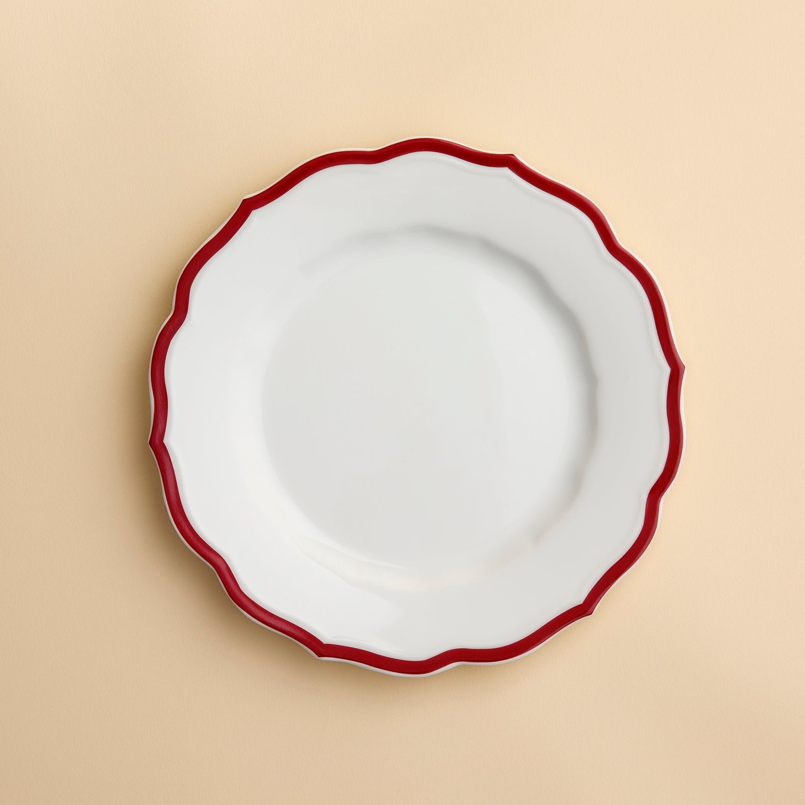The Stella Scalloped Crimson Salad Plate by Caskata, featuring a white ceramic surface with a wavy crimson rim inspired by vintage dinnerware, is displayed on a light beige background.