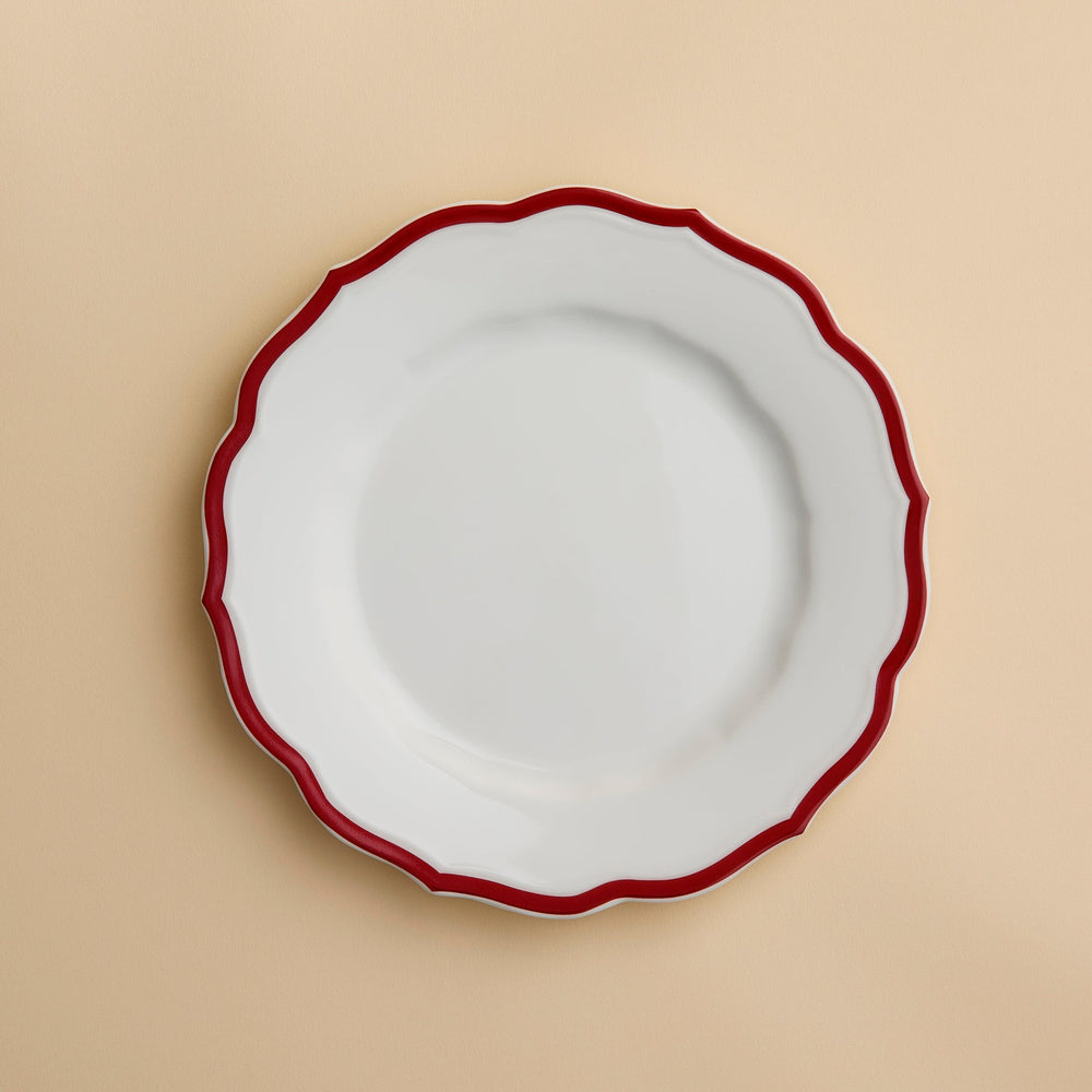 The Stella Scalloped Crimson Salad Plate by Caskata, featuring a white ceramic surface with a wavy crimson rim inspired by vintage dinnerware, is displayed on a light beige background.