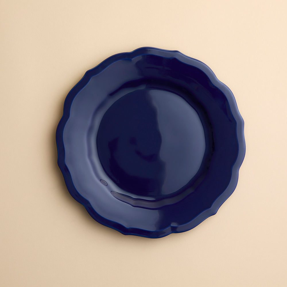 A  dark blue Caskata Stella Scalloped Solid Blue Salad Plate with a scalloped edge in porcelain on a creamy beige background.