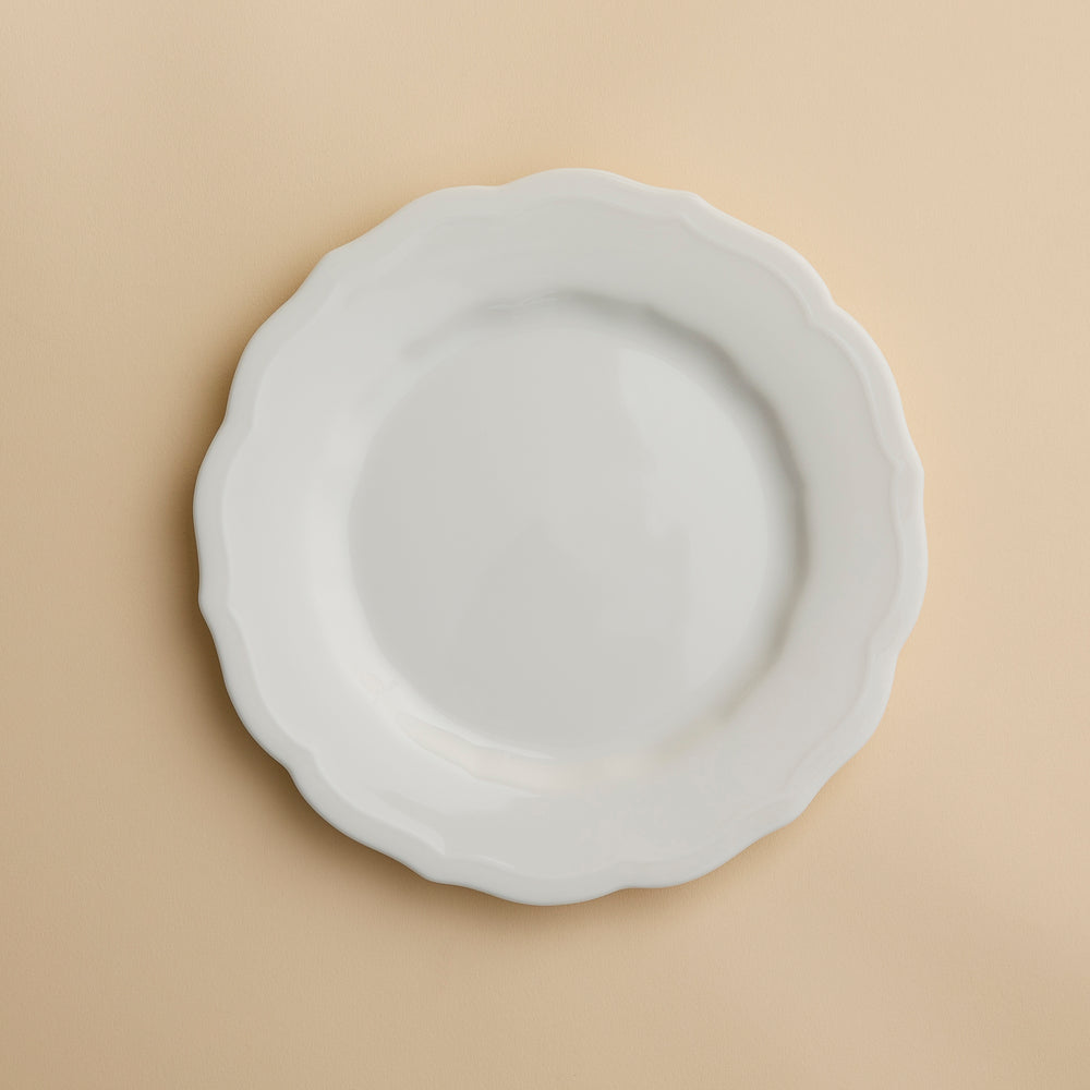 The Stella Scalloped White Salad Plate by Caskata, a white, round handcrafted plate with a scalloped edge, is centered on a beige background.