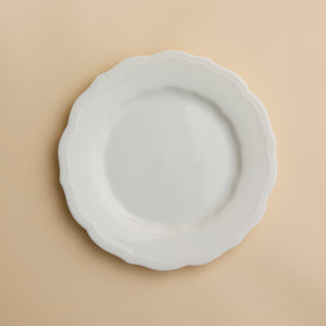 The Stella Scalloped White Salad Plate by Caskata, a white, round handcrafted plate with a scalloped edge, is centered on a beige background.