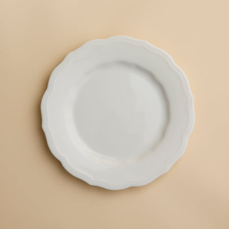 The Stella Scalloped White Salad Plate by Caskata, a white, round handcrafted plate with a scalloped edge, is centered on a beige background.