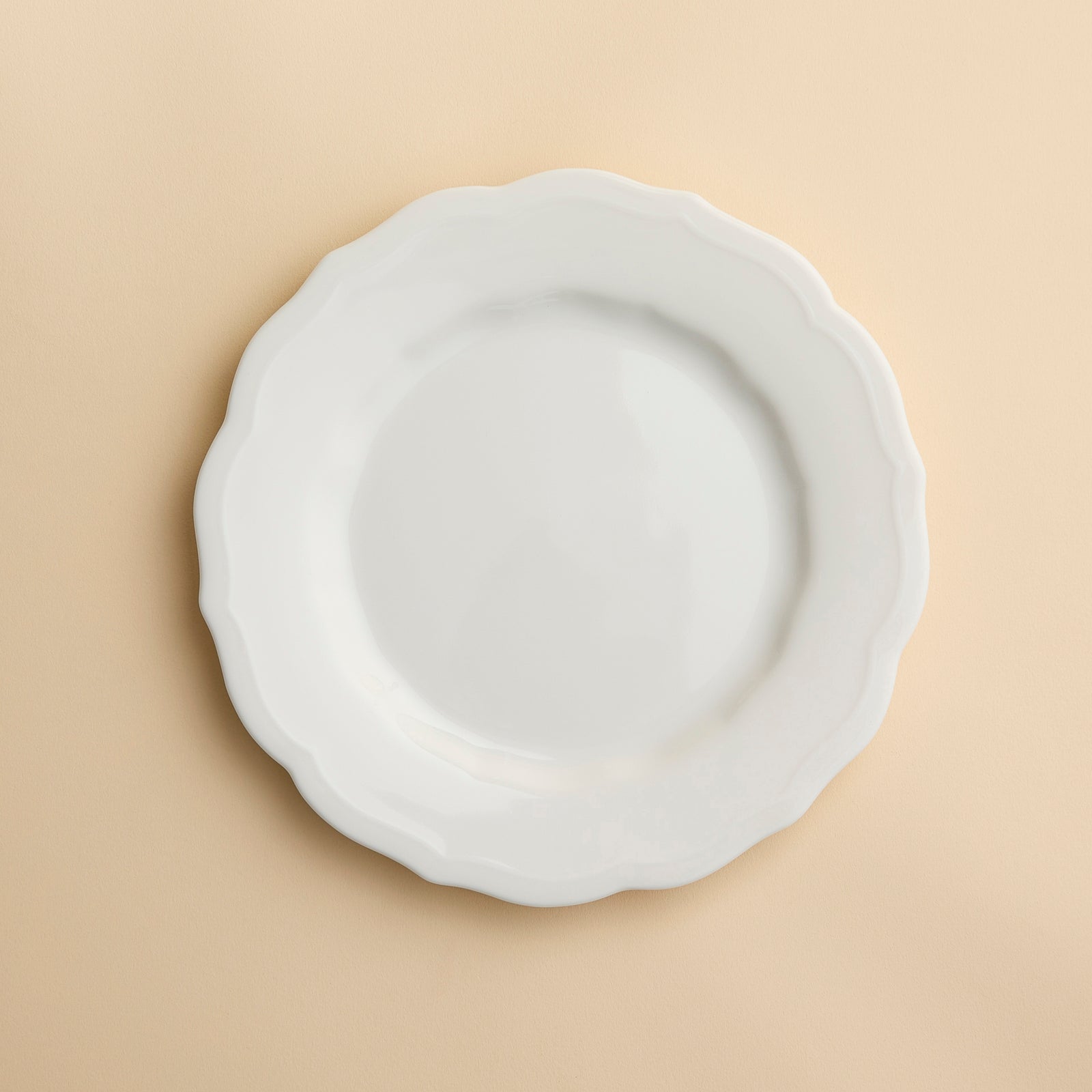 The Stella Scalloped White Salad Plate by Caskata, a white, round handcrafted plate with a scalloped edge, is centered on a beige background.