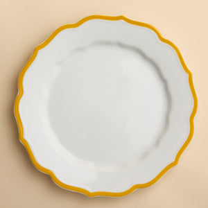 The Stella Scalloped Marigold Dinner Plate by Caskata features a white porcelain design with a decorative scalloped edge and yellow border, shown from above on a creamy beige background.