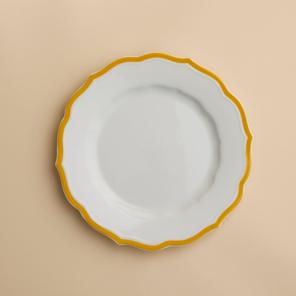 The Stella Scalloped Marigold Salad Plate by Caskata features a round white ceramic design with a wavy yellow rim and sits elegantly on a beige background.