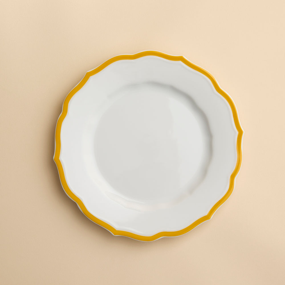 The Stella Scalloped Marigold Salad Plate by Caskata features a round white ceramic design with a wavy yellow rim and sits elegantly on a beige background.