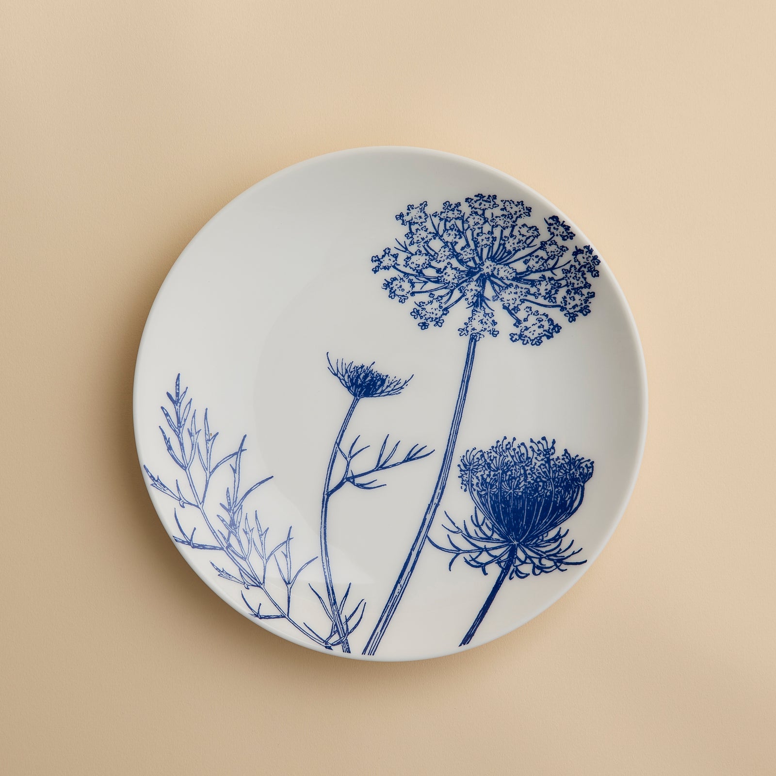 White ceramic plate adorned with blue botanical illustrations of flower stems and leaves, set on a beige background. The Summer Blues Coupe Salad Plate by Caskata Artisanal Home brings a sophisticated botanical touch to your porcelain dinnerware collection.