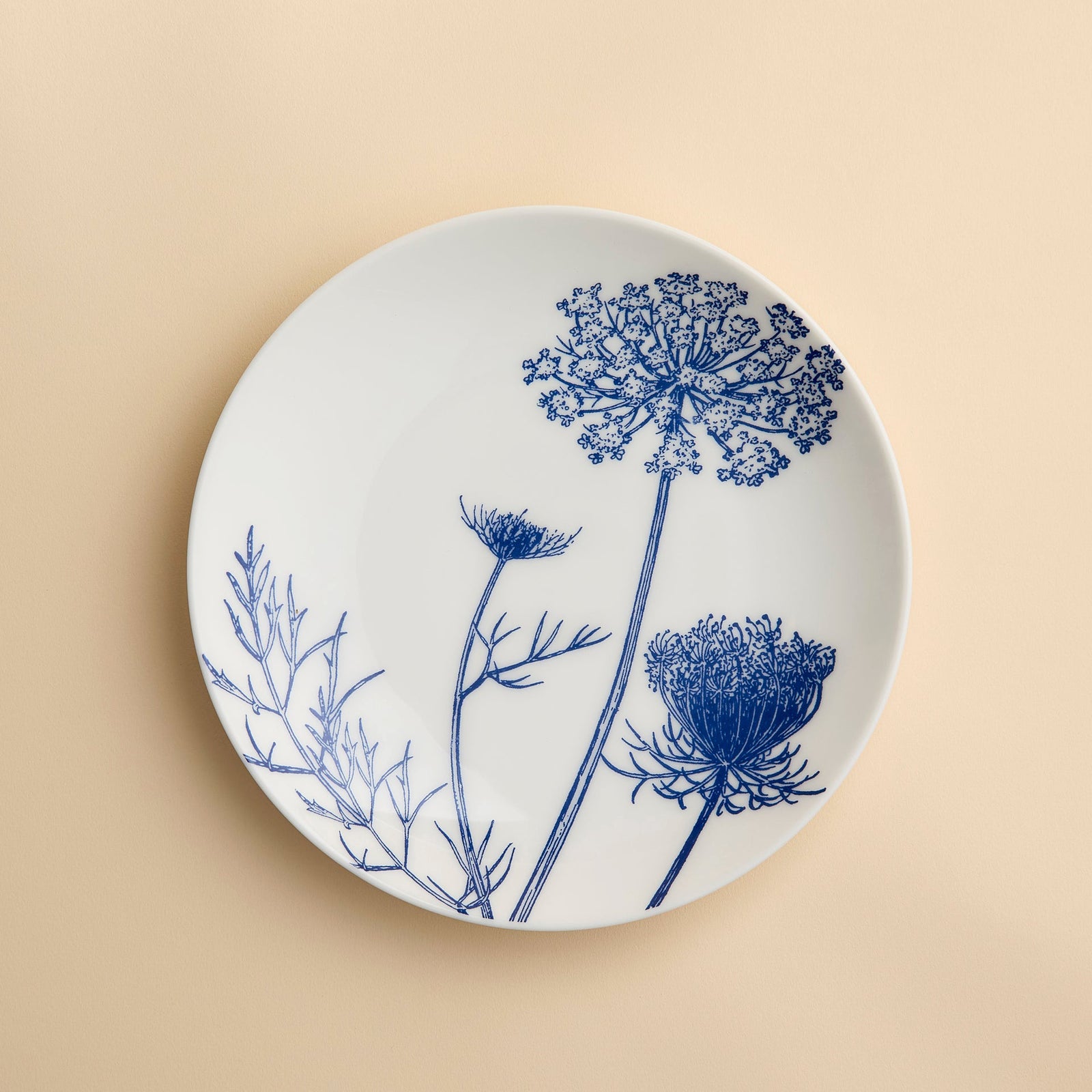 White ceramic plate adorned with blue botanical illustrations of flower stems and leaves, set on a beige background. The Summer Blues Coupe Salad Plate by Caskata Artisanal Home brings a sophisticated botanical touch to your porcelain dinnerware collection.