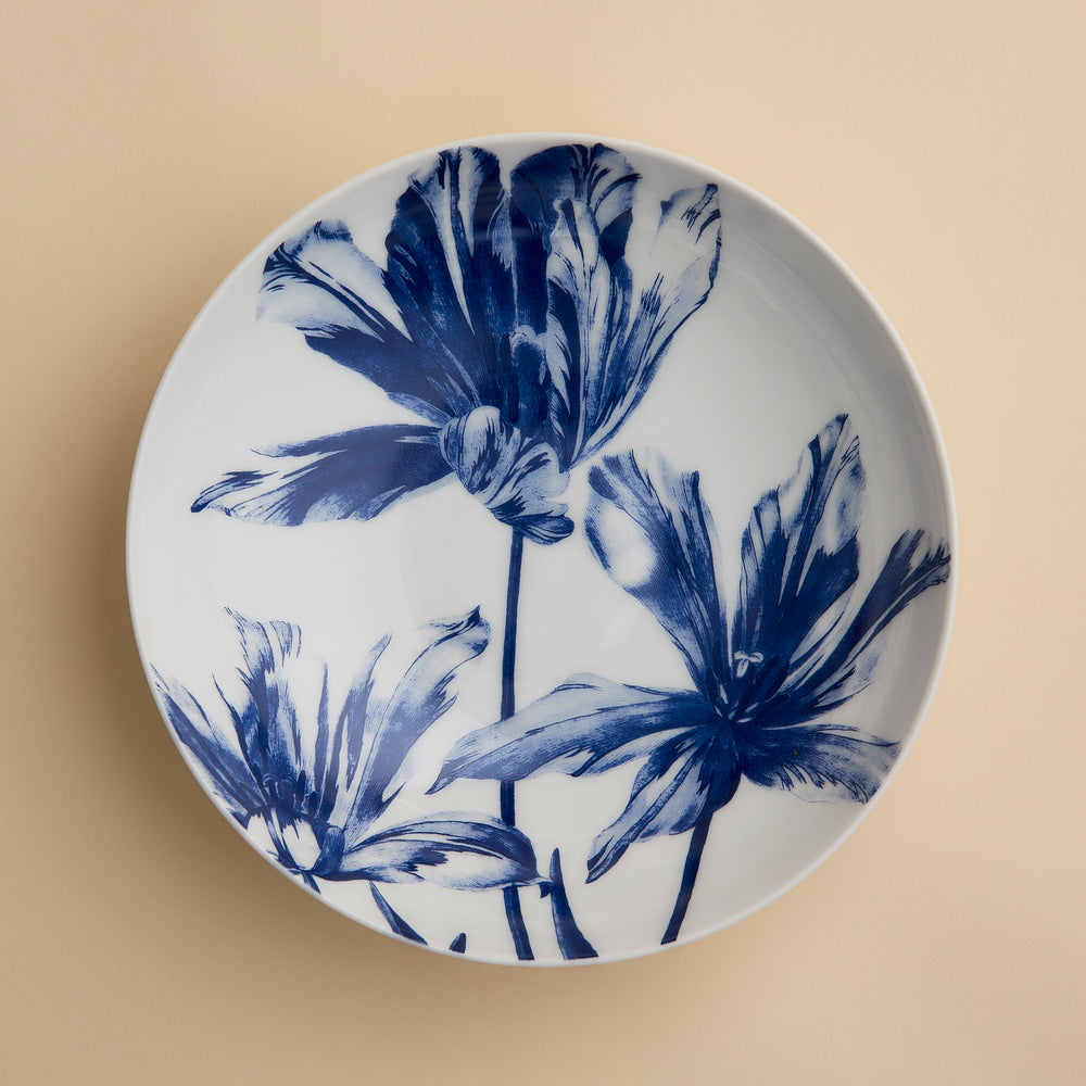 The Caskata Tulip Entrée Bowl is a white ceramic plate with a beige background, adorned with three large abstract blue flowers—perfect as a stylish centerpiece or for serving as an elegant blue floral bowl.