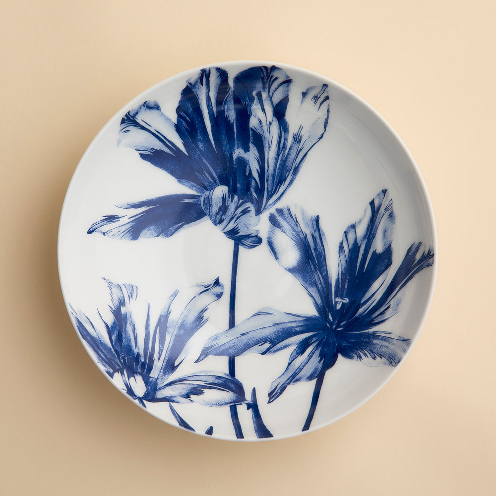The Caskata Tulip Entrée Bowl is a white ceramic plate with a beige background, adorned with three large abstract blue flowers—perfect as a stylish centerpiece or for serving as an elegant blue floral bowl.