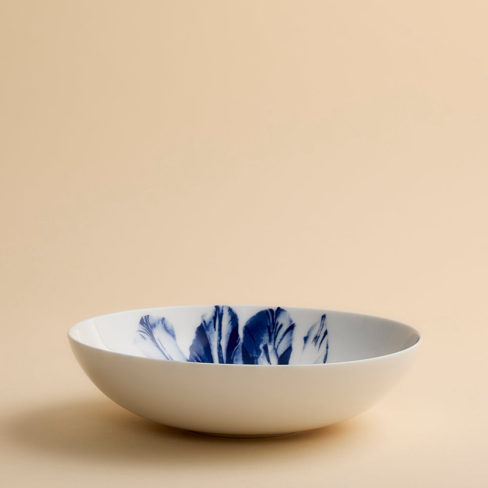 A shallow white Caskata Tulip Entrée Bowl featuring delicate blue leaf-like patterns on the interior, displayed against a plain beige background.