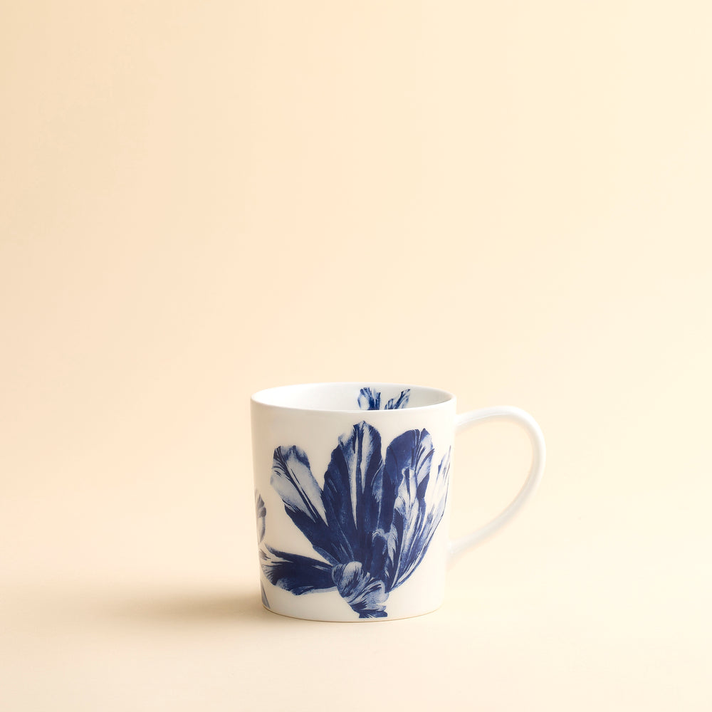 The Caskata Tulip Mug features a delicate blue botanical floral design on white high-fired porcelain, combining elegance and durability. Perfect for your favorite beverages, this mug is shown on a light beige surface with a matching neutral background.