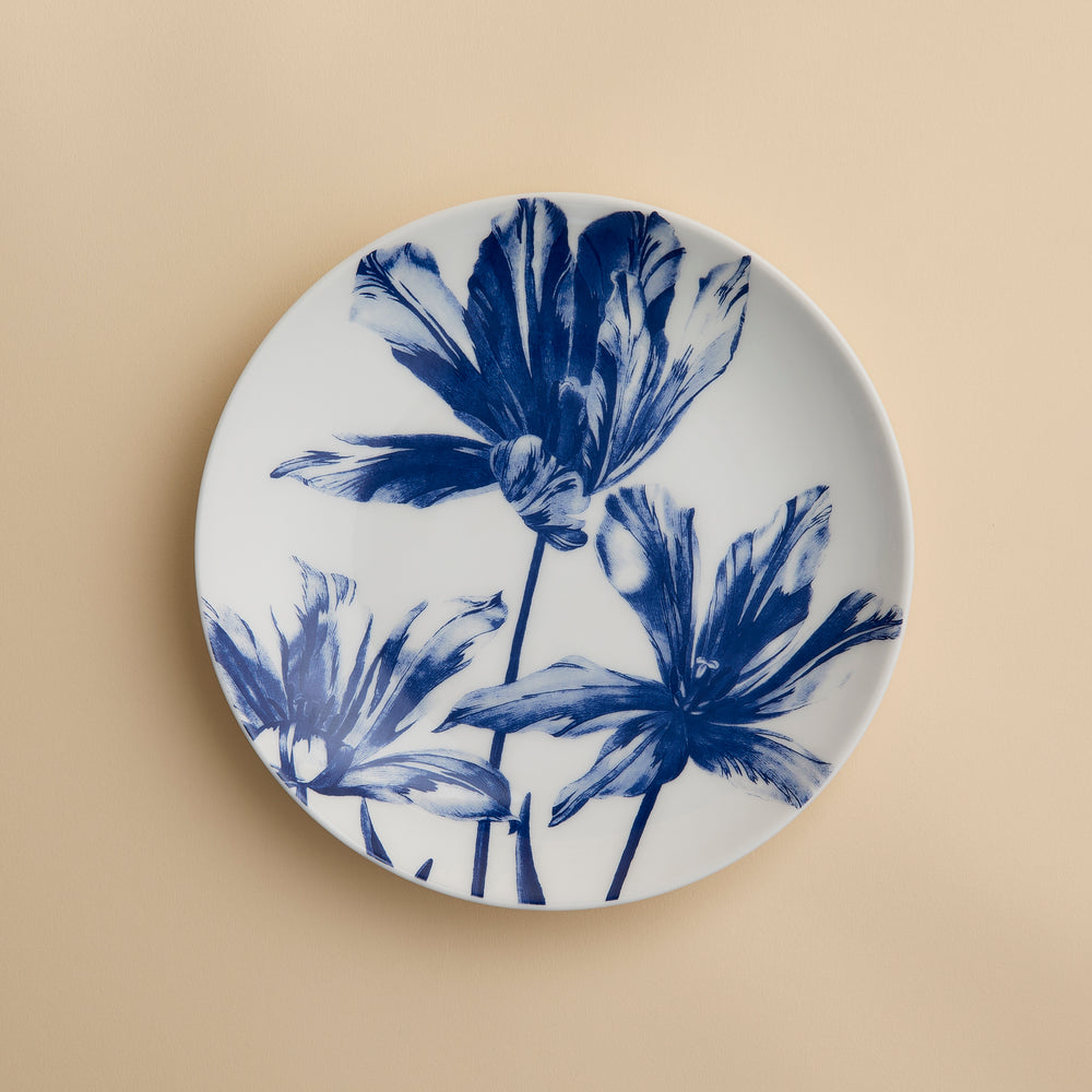 The Caskata Tulip Coupe Salad Plate is a white ceramic plate adorned with a blue floral design featuring three large flowers, beautifully displayed against a plain beige background.