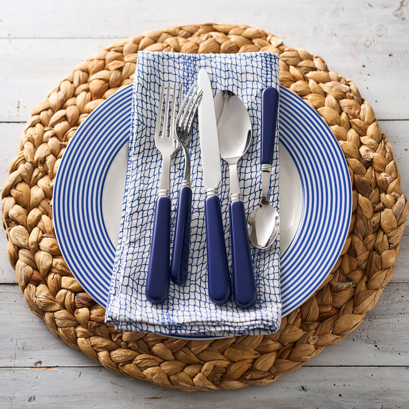 A place setting with blue-handled cutlery arranged on a napkin, placed on a Newport Stripe Rimmed Dinner Plate by Caskata Artisanal Home, all atop a braided wicker placemat.