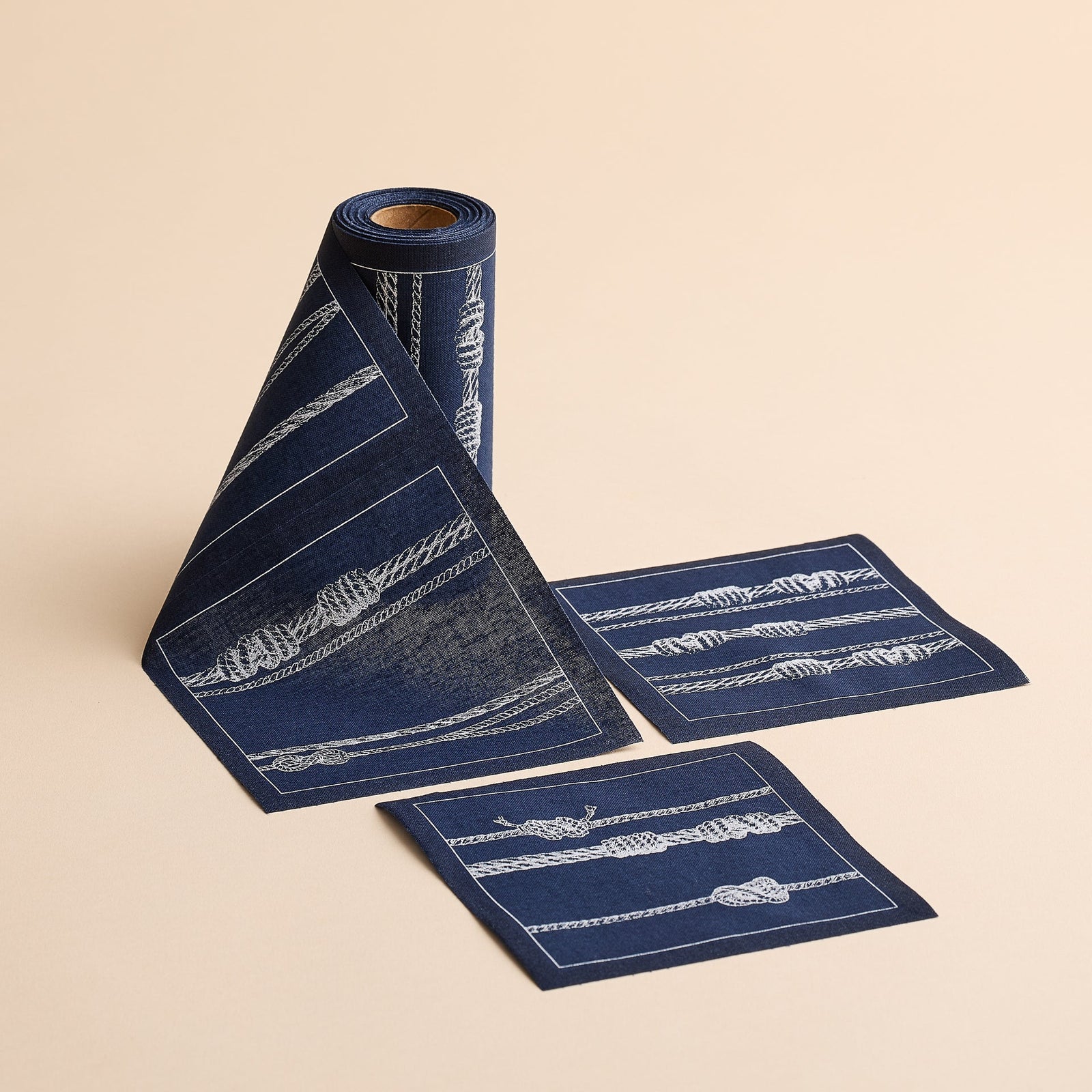 A partially unrolled MY DRAP Sailors Knots Cocktail Napkin Roll—navy blue with white rope designs—rests on a beige background, accompanied by two matching square napkins, ideal as reusable party or cotton cocktail napkins.
