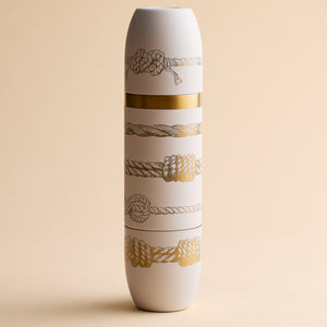 Introducing the Caskata x High Camp Crema Sailors Knots Wine Flask by Caskata—a tall, white insulated flask adorned with gold and black rope patterns inspired by nautical motifs and accented with a striking gold band at its center, beautifully presented against a beige background. This stylish wine flask blends maritime charm with sophisticated design.