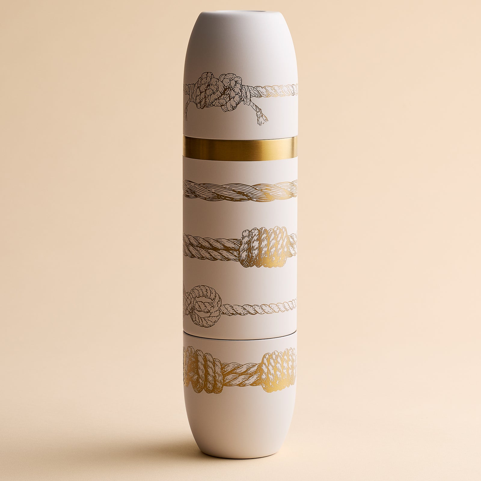 Introducing the Caskata x High Camp Crema Sailors Knots Wine Flask by Caskata—a tall, white insulated flask adorned with gold and black rope patterns inspired by nautical motifs and accented with a striking gold band at its center, beautifully presented against a beige background. This stylish wine flask blends maritime charm with sophisticated design.