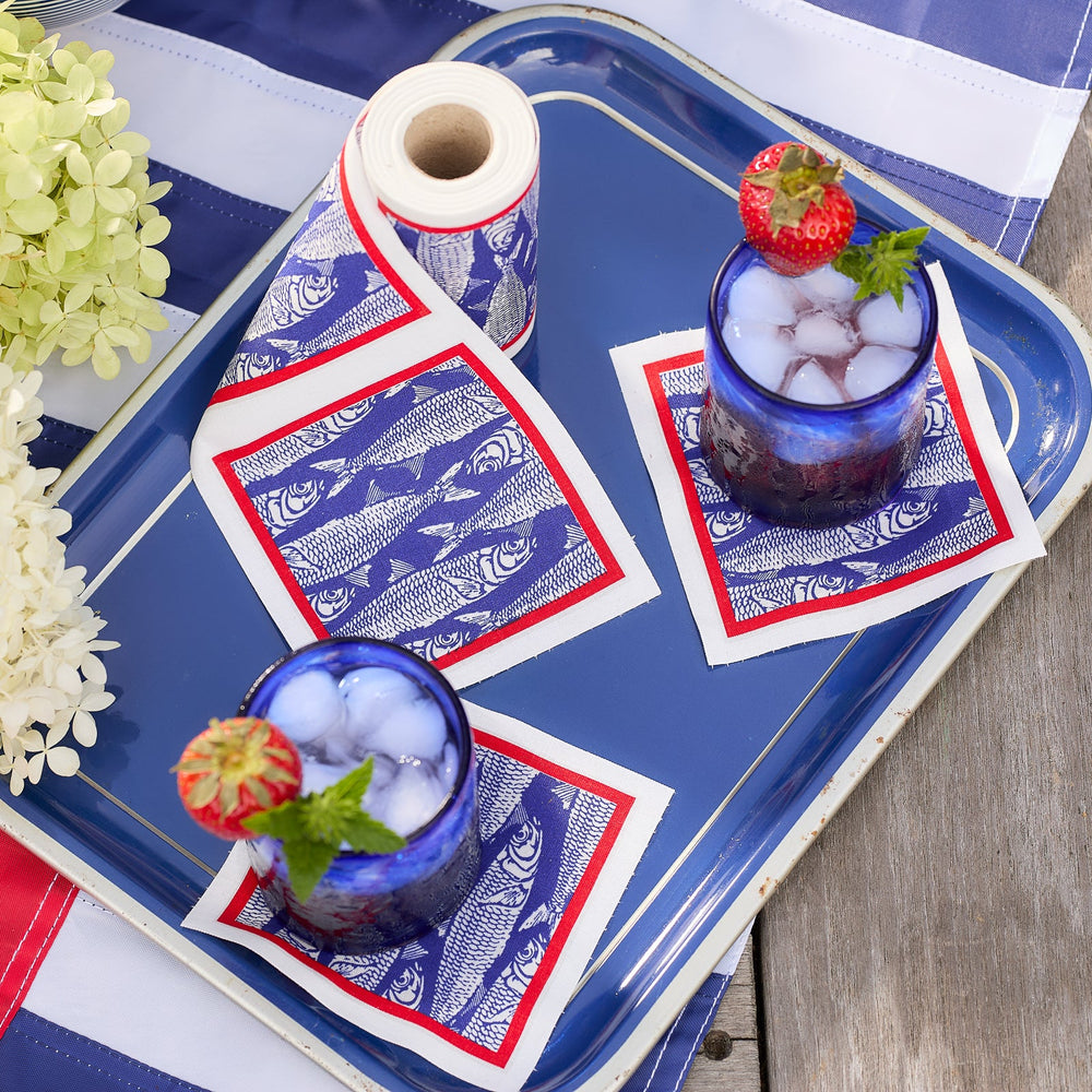 Two blue glasses filled with iced drinks and garnished with strawberries sit on a blue tray, accented by MY DRAP Sardines Cocktail Napkin Roll cotton cocktail napkins in a blue, white, and red block print fish pattern, alongside white flowers.