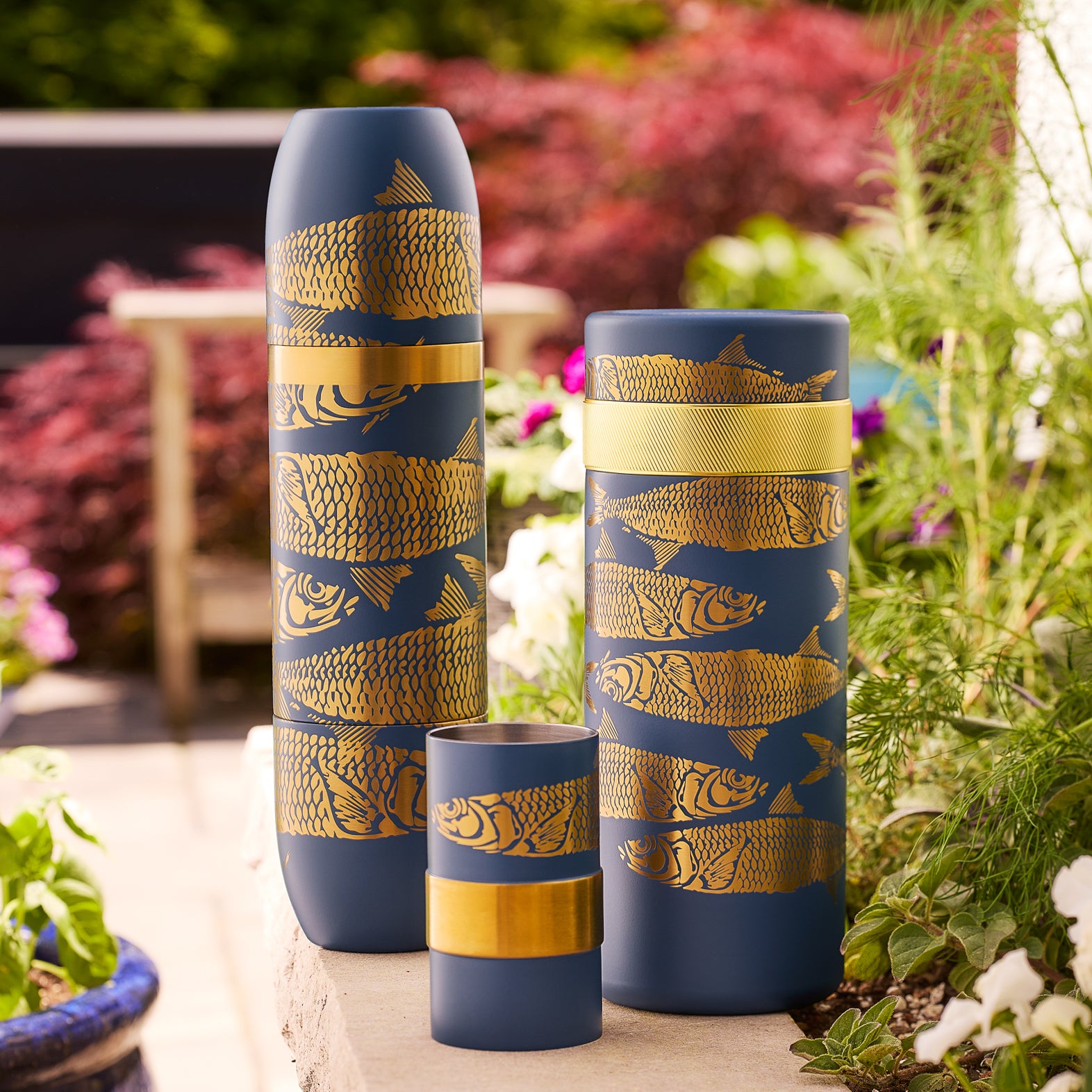 Three matte blue vases with gold fish and scale patterns from Caskata are displayed outdoors with greenery—chic accents to pair with the Caskata X High Camp Sardines Bar Master Bundle for your next wine or cocktail gathering.