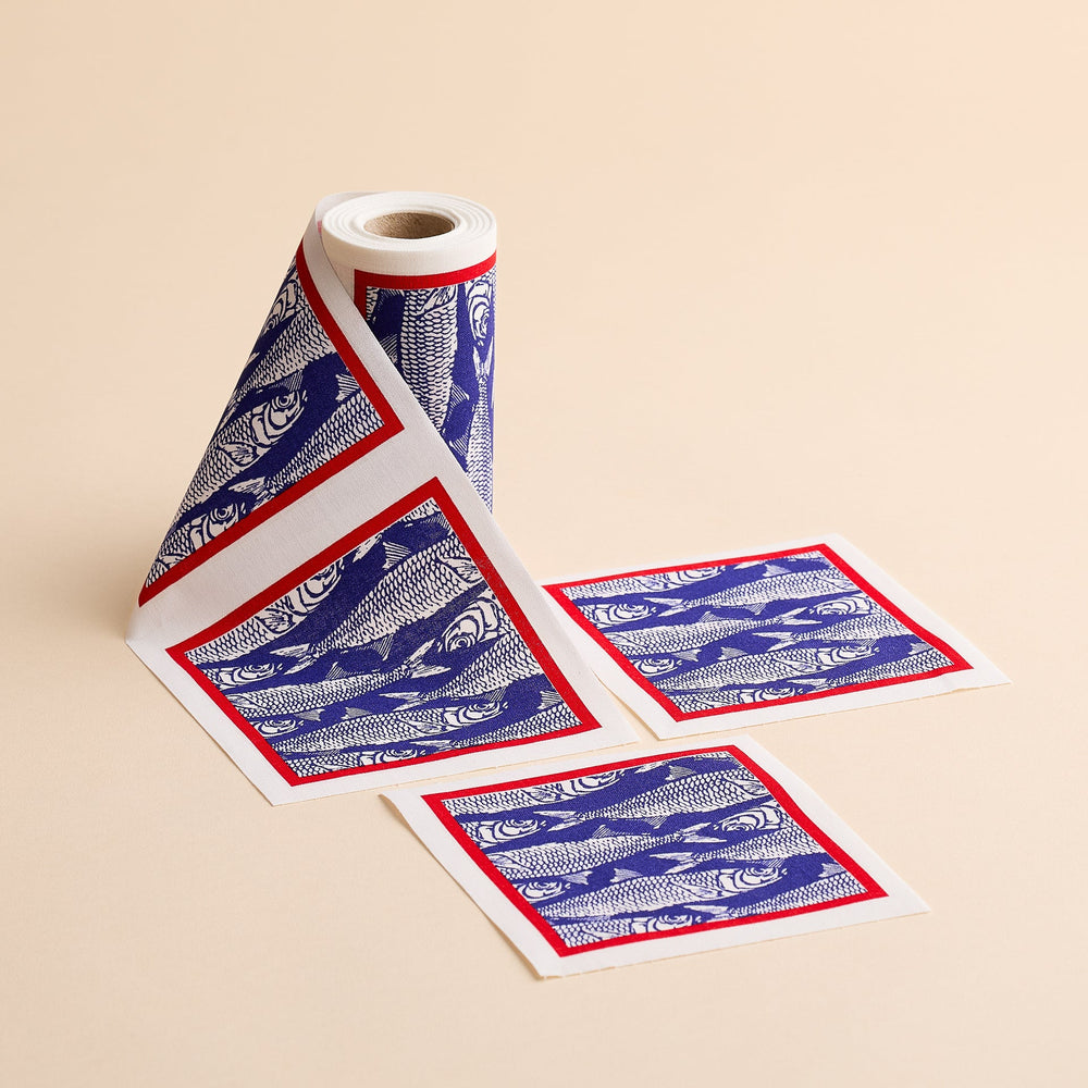 A roll of patterned paper from MY DRAP, the Sardines Cocktail Napkin Roll, showcases a charming block print fish design with blue fish illustrations and red borders. Two individual square sheets—resembling cotton cocktail napkins—have been torn off and are placed beside the roll on a beige surface.