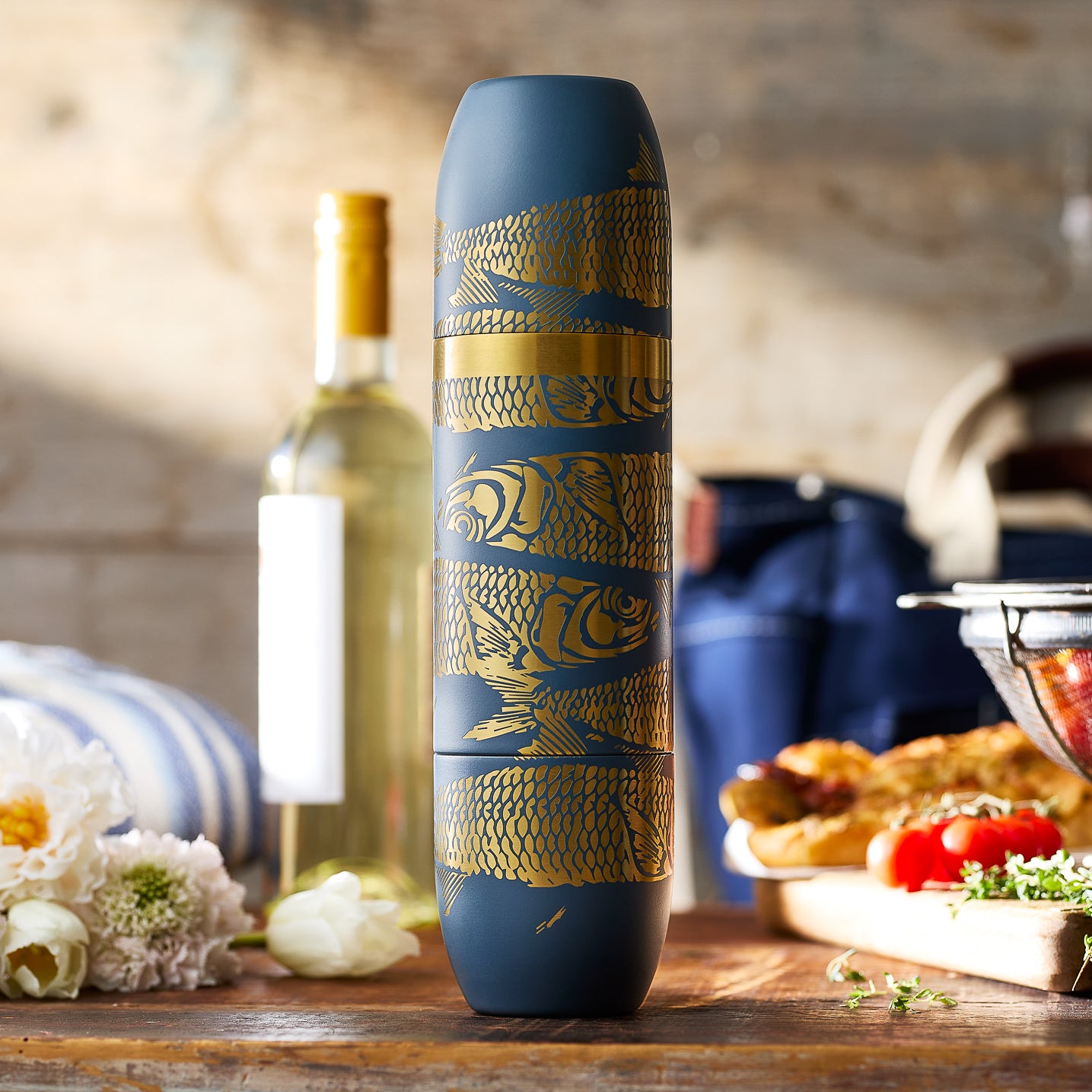 The Caskata x High Camp Dark Blue Sardines Parkside Flask features gold fish and scale designs and comes with a gray felt carrying case, blending coastal-inspired style with the convenience of magnetized cups.