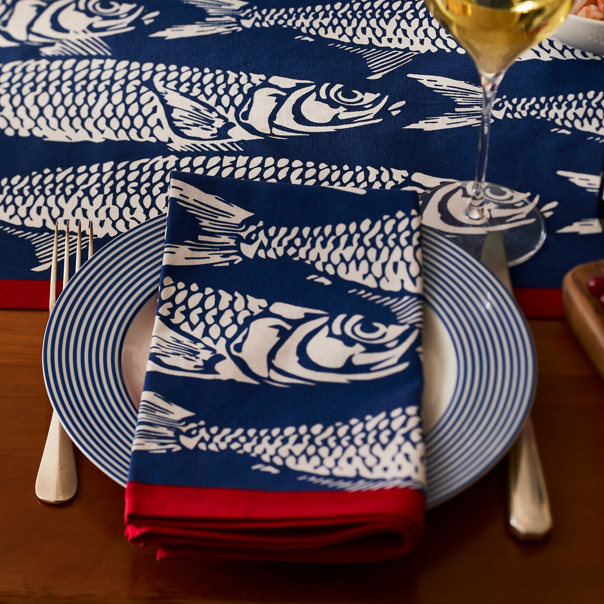 A plate with a blue and white fish-patterned napkin from Caskata’s Sardines Dinner Napkins, Set of 4, is set on a table with cutlery and a glass of white wine—the perfect stylish touch for your dining experience.