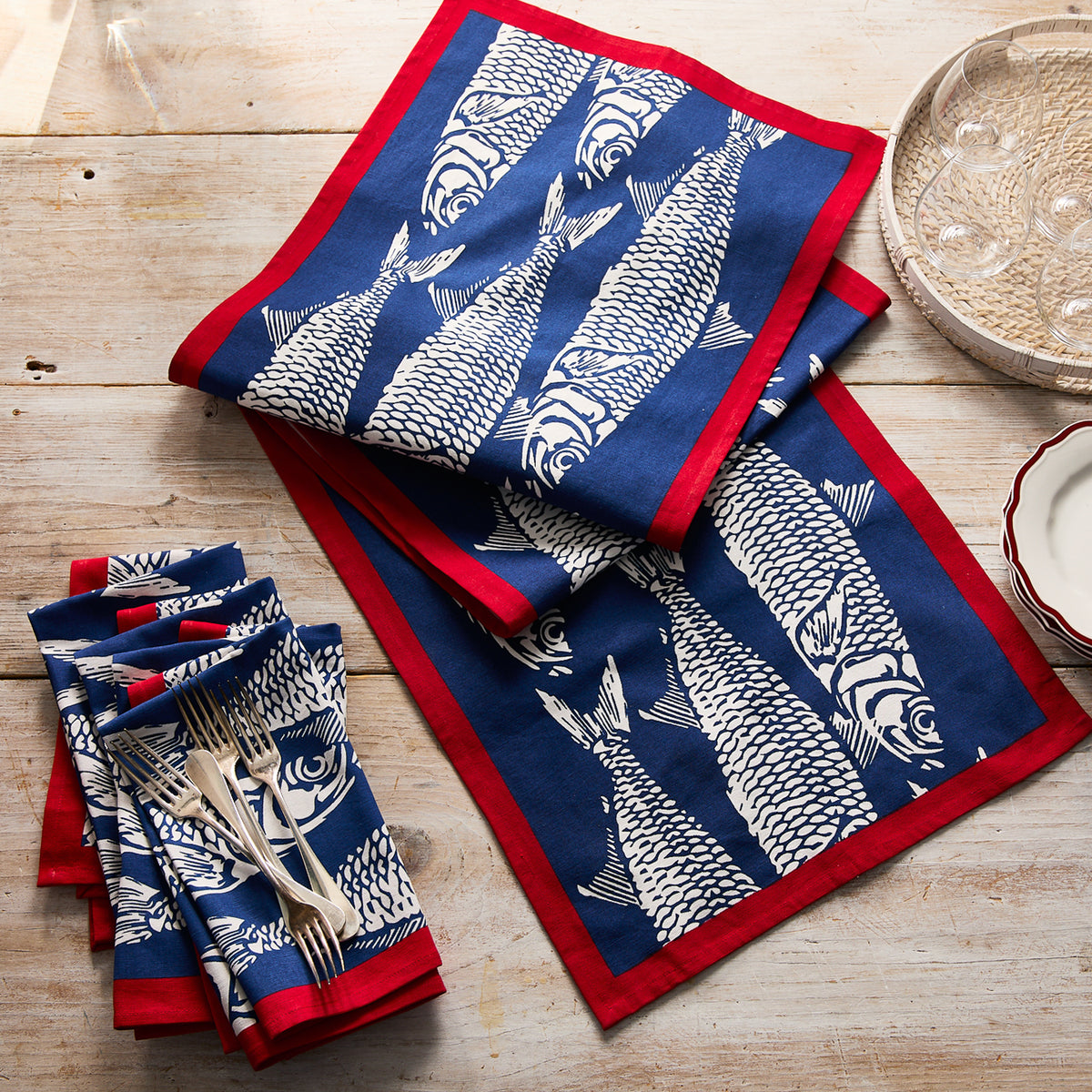 Blue and white napkins with fish motifs and red borders add seaside charm to a wooden table, beautifully set with stacked plates, silverware, and the Sardines Table Runner by Caskata.