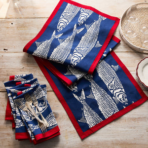 Blue and white napkins with fish motifs and red borders add seaside charm to a wooden table, beautifully set with stacked plates, silverware, and the Sardines Table Runner by Caskata.