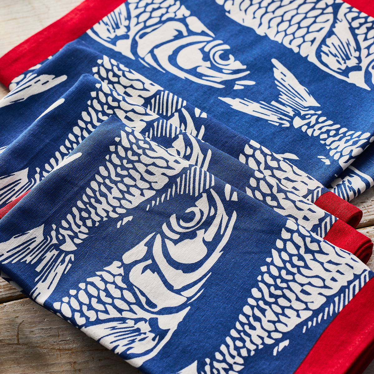 Caskata Sardines Dinner Napkins, set of 4, feature blue and red cotton napkins with white sardine prints arranged on a wooden surface.