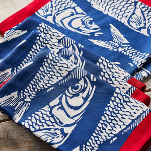 Caskata Sardines Dinner Napkins, set of 4, feature blue and red cotton napkins with white sardine prints arranged on a wooden surface.