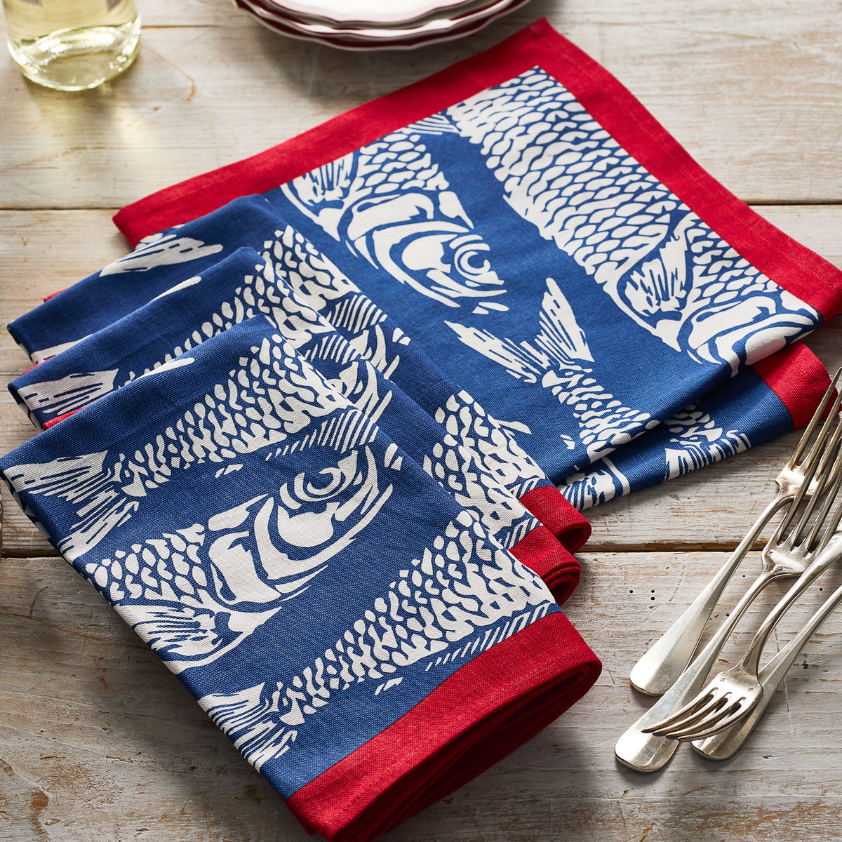 The Caskata Sardines Dinner Napkins, Set of 4, feature a blue and white block print sardines design with red borders and are neatly folded on a wooden table beside plates, cutlery, and a glass.