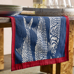 The Caskata Sardines Table Runner, featuring a white fish pattern and red border on cotton, brings seaside charm to your table as it drapes across a wooden surface set with plates, glasses, and wine.