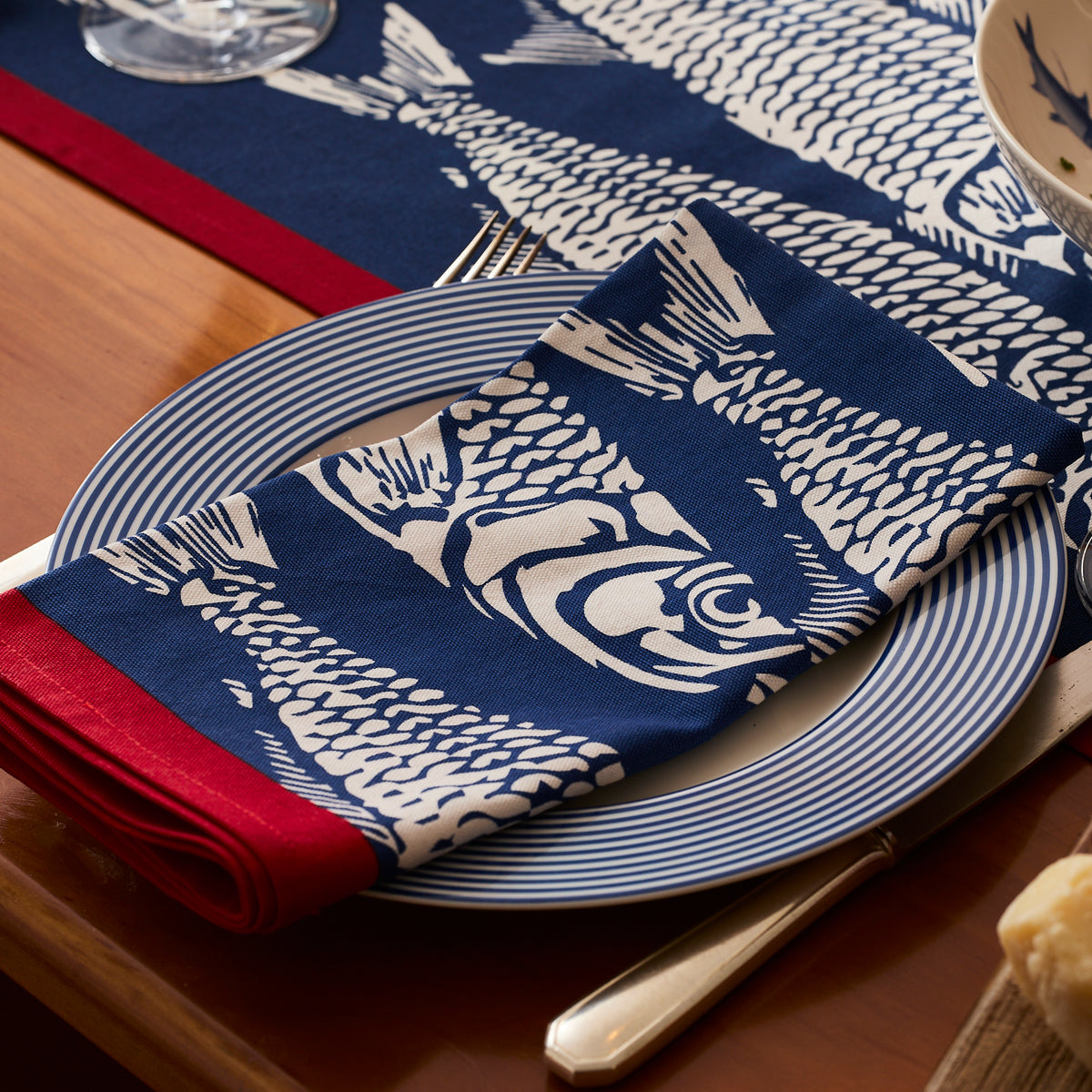 A Caskata Sardines Dinner Napkin from the set of 4 is folded on a striped plate, placed on a wooden table with cutlery and a coordinating table runner.