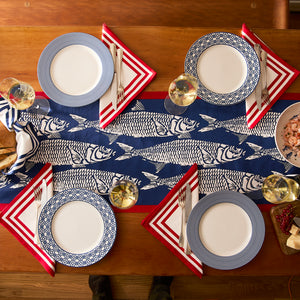 A wooden table set for four with a blue and white fish-patterned runner, striped plates, Caskata Triple Stripe Red Dinner Napkins, wine glasses, bread, and a bowl of seafood creates a charming coastal dining scene.