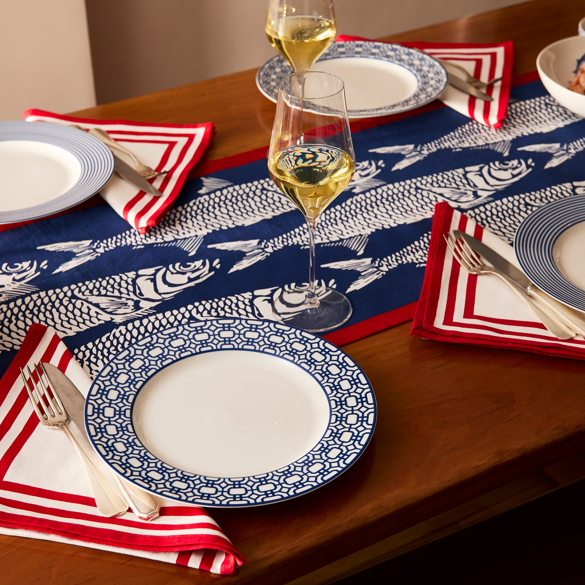 Create a charming seaside table decor with Caskata's Sardines Table Runner, wooden dining set, blue and white patterned plates, red and white napkins, silver cutlery, and glasses of white wine.