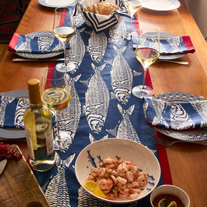 A Sardines Table Runner by Caskata, paired with fish-patterned blue and white linens, plates, wine glasses, shrimp, white wine, bread, and red napkins creates charming seaside-inspired dining decor.