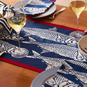 A seaside table is set with Caskata's Sardines Table Runner, matching blue and white napkins, striped plates, wine glasses, and coastal decor on a wooden surface.