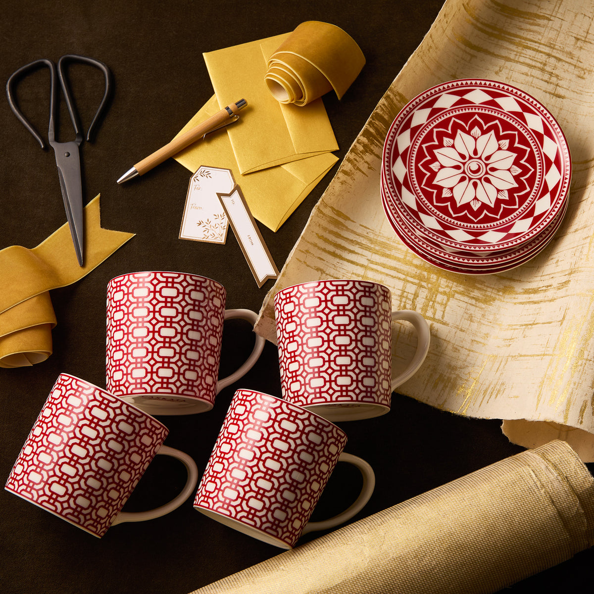 The "Scandi Holiday" Bundle by Caskata features four red and white porcelain mugs and three small plates, arranged with yellow paper, scissors, a pen, and gift tags on a dark surface. All items are microwave safe.