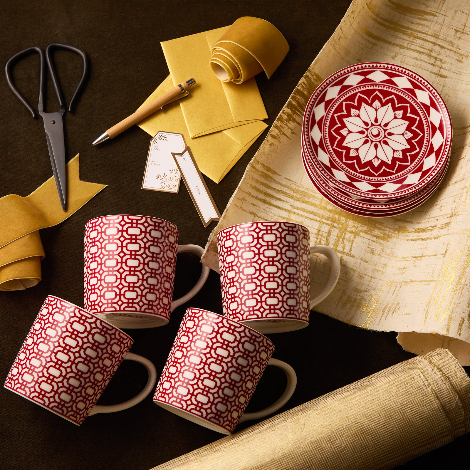 The "Scandi Holiday" Bundle by Caskata features four red and white porcelain mugs and three small plates, arranged with yellow paper, scissors, a pen, and gift tags on a dark surface. All items are microwave safe.