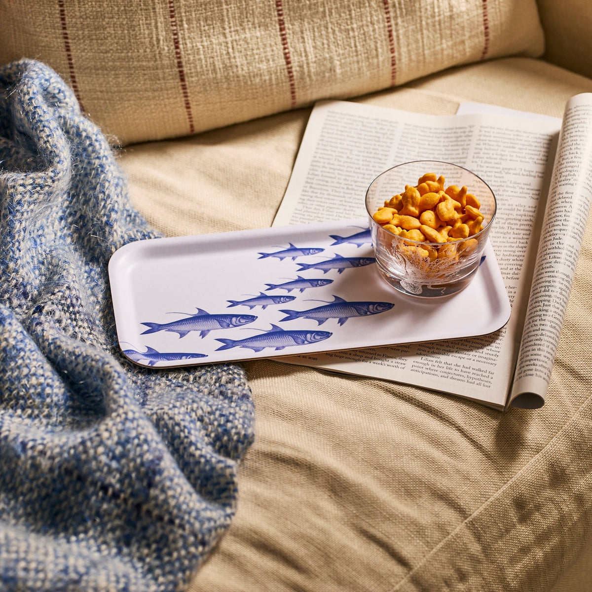 A glass bowl of goldfish crackers rests on Caskata’s School of Fish Small Rectangular Birchwood Tray atop an open magazine, with a blue knit blanket draped over a beige sofa.