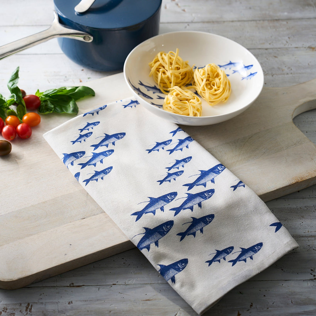 A kitchen scene with white dinnerware collection including a bowl containing pasta on a School of Fish Kitchen Towel, Set of 2 by Caskata, next to fresh basil and tomatoes, with a blue pot and tongs in the background.