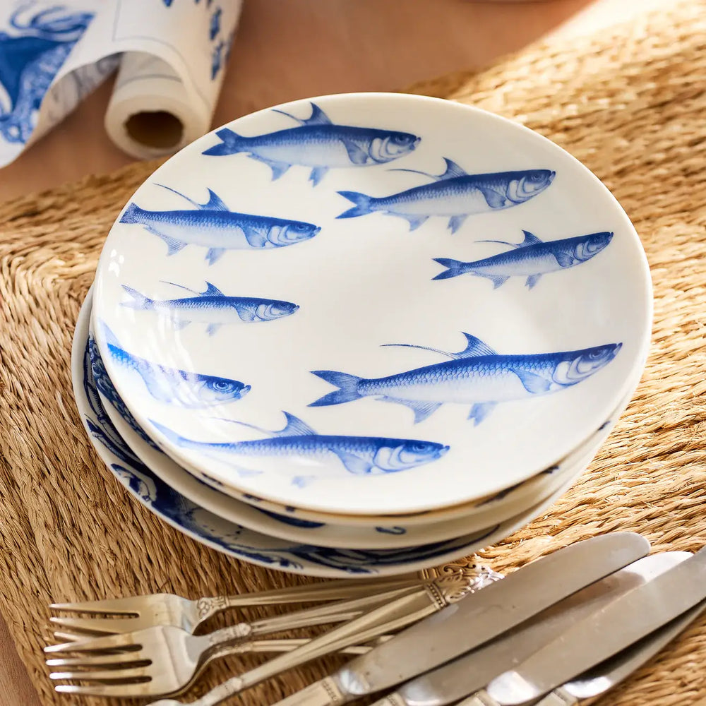 A stack of Caskata Coastal Icons Small Plates, Mixed Set of 4—crafted from white porcelain and decorated with blue fish motifs—rests on a woven placemat next to silver forks and knives.